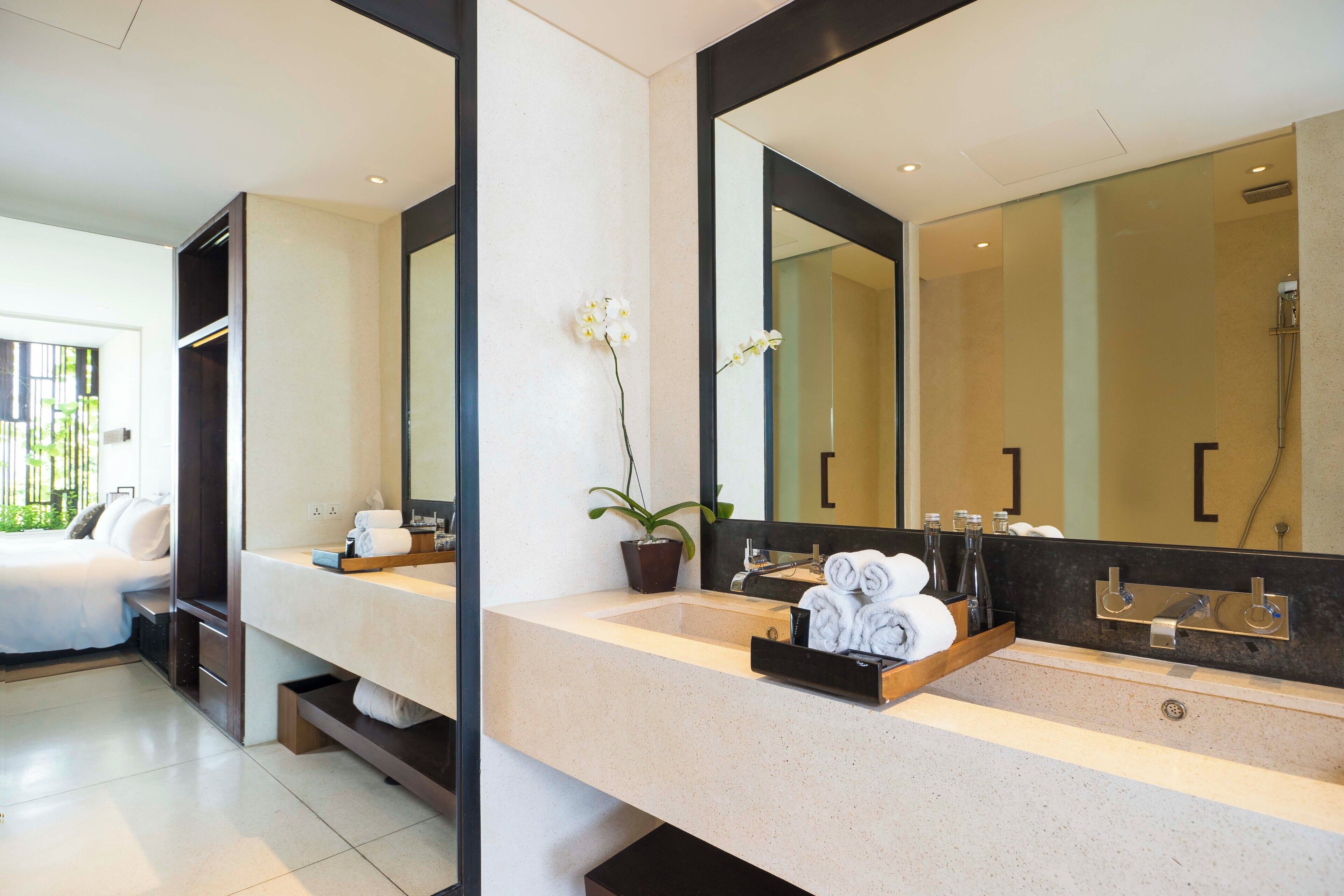 A modern bathroom with a large mirror, a sink, and a shower. There is a potted plant and a tray with towels on the counter.