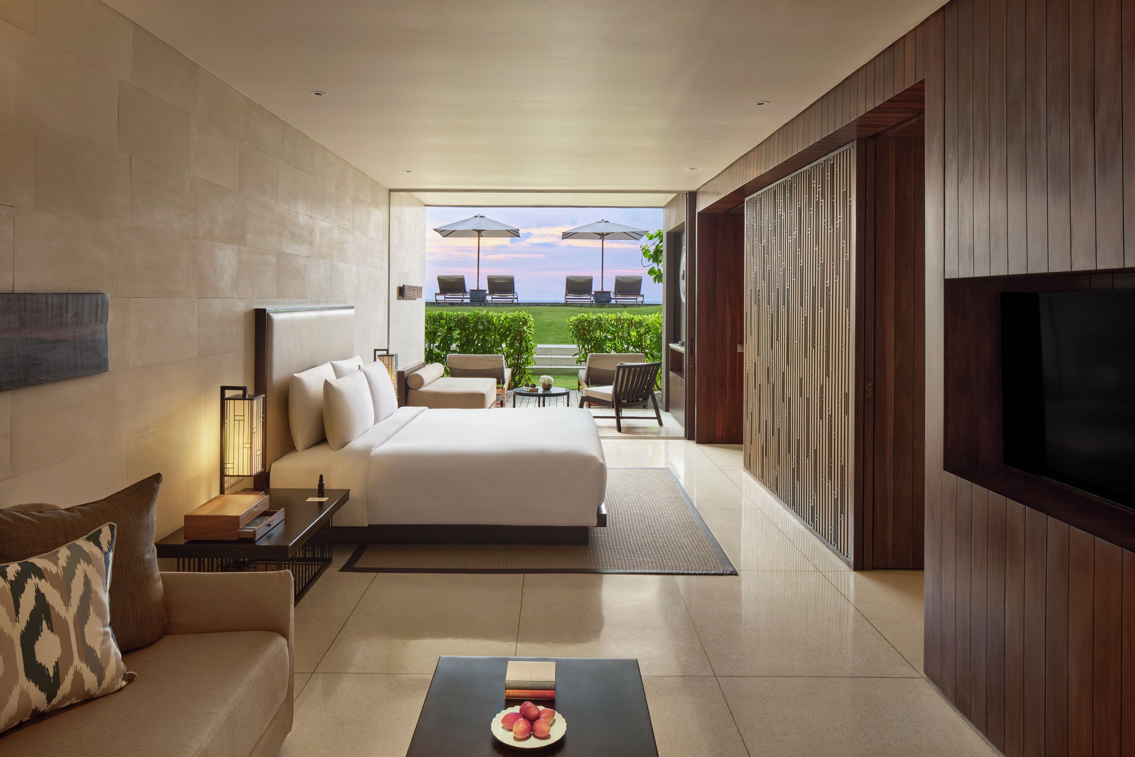 A modern hotel room with a large bed, a sofa, a coffee table, and a TV. There is a balcony with outdoor seating and a view of the sea.