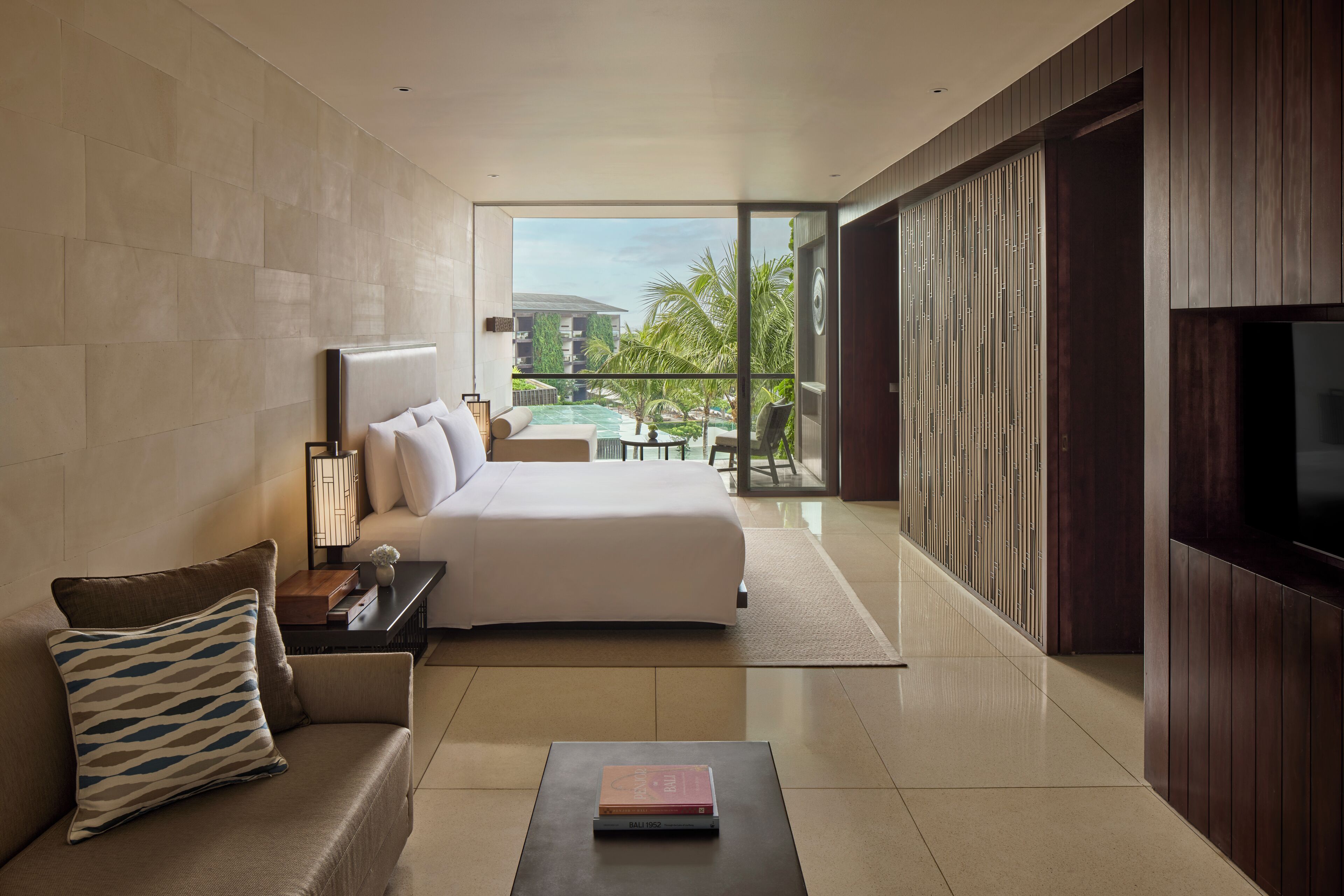 A modern hotel room with a large bed, a sofa, a coffee table, and a TV. The room has a view of a pool and palm trees.