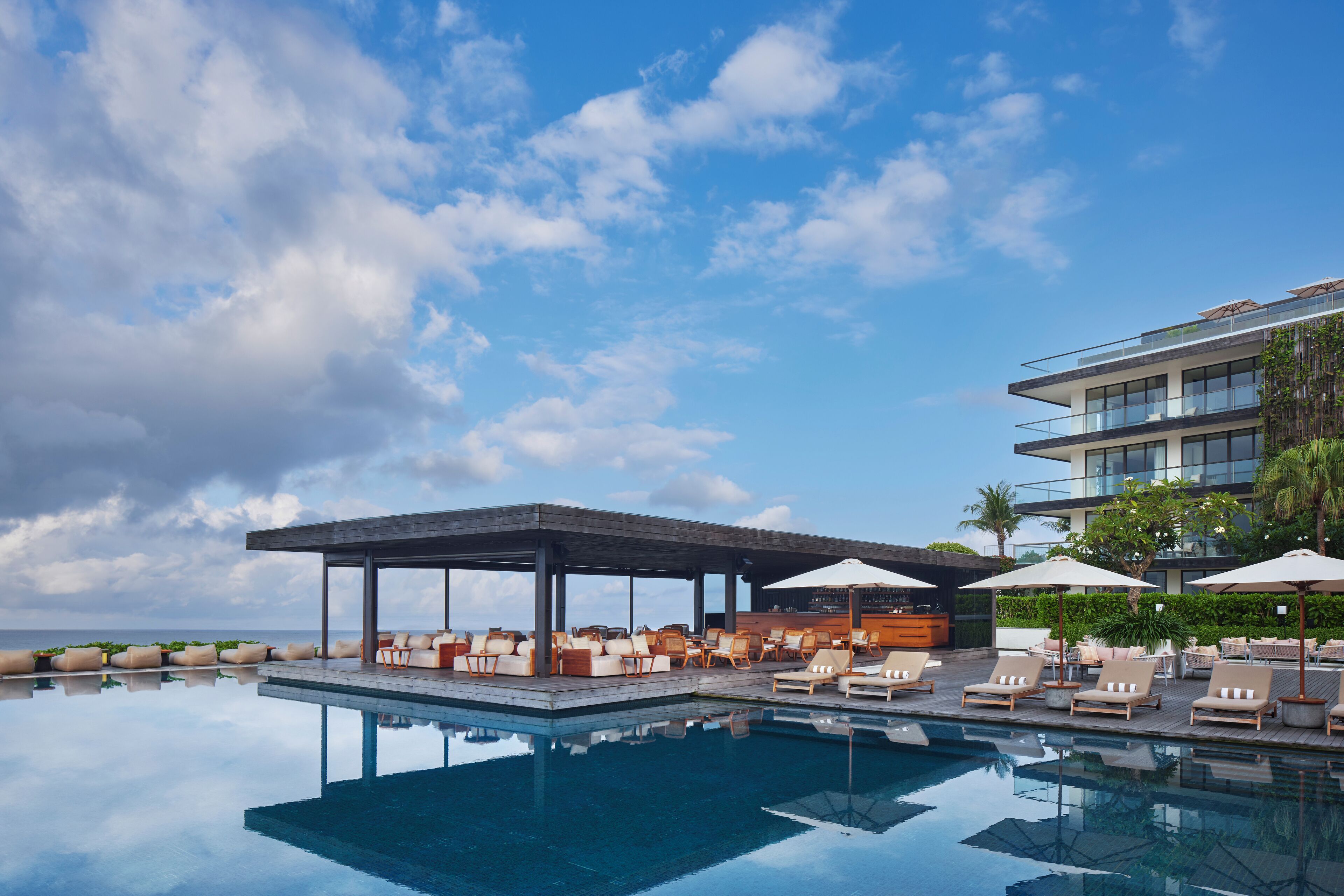A pool area with lounge chairs and umbrellas, a covered seating area, and a modern building with balconies.