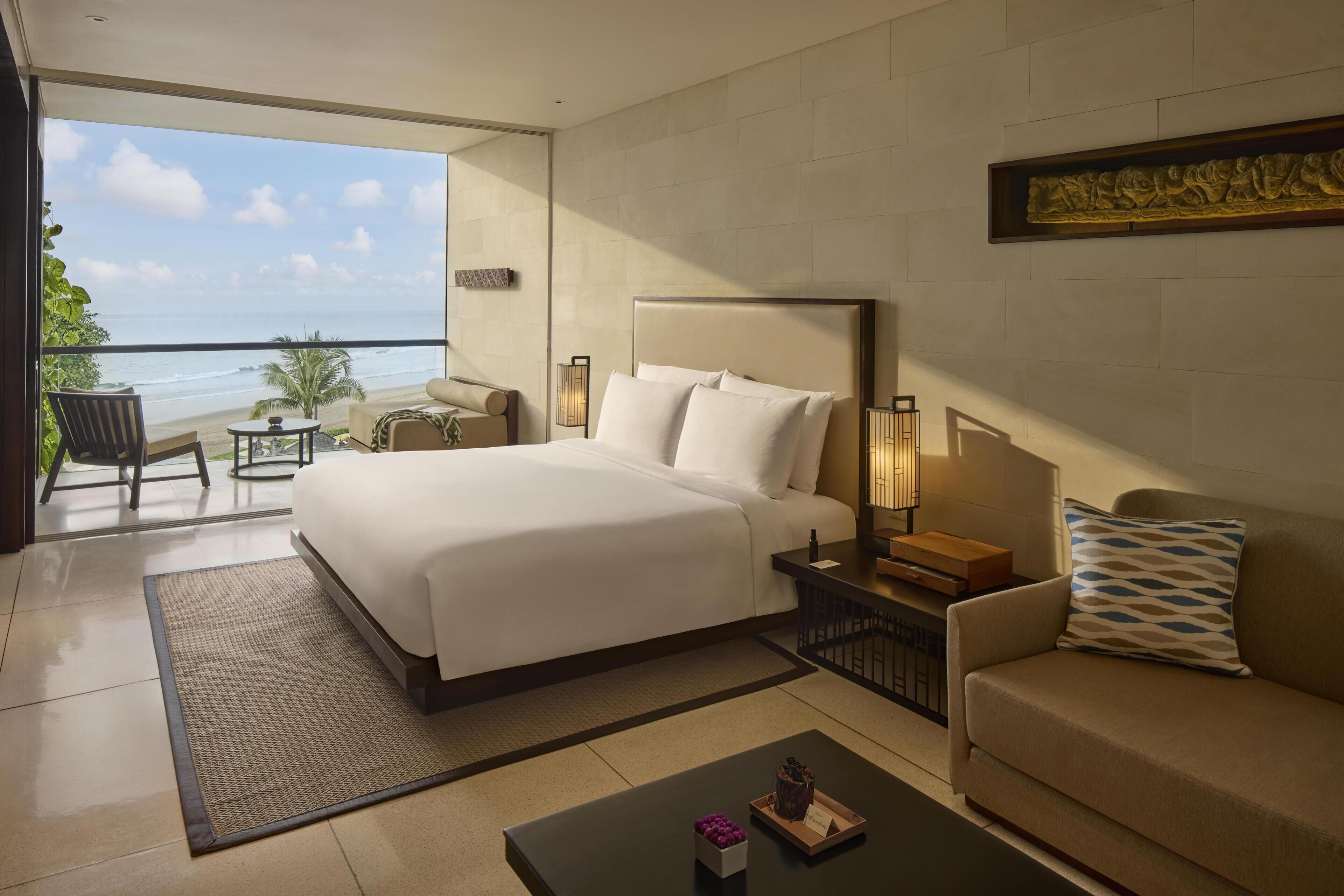 A hotel room with a bed, a sofa, a coffee table, a nightstand, and a view of the beach.