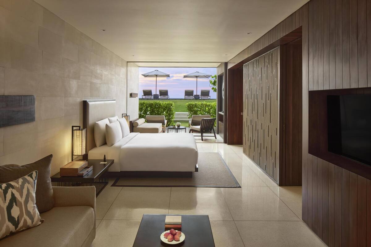 A modern hotel room with a bed, a sofa, a coffee table, a TV, and a view of the outdoors.