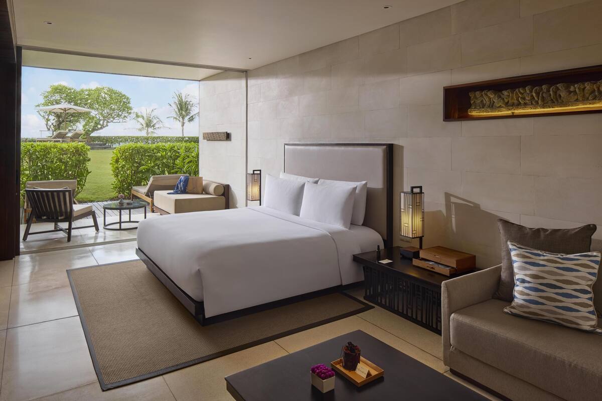 A modern hotel room with a large bed, a sofa, a coffee table, and a view of the outdoors.