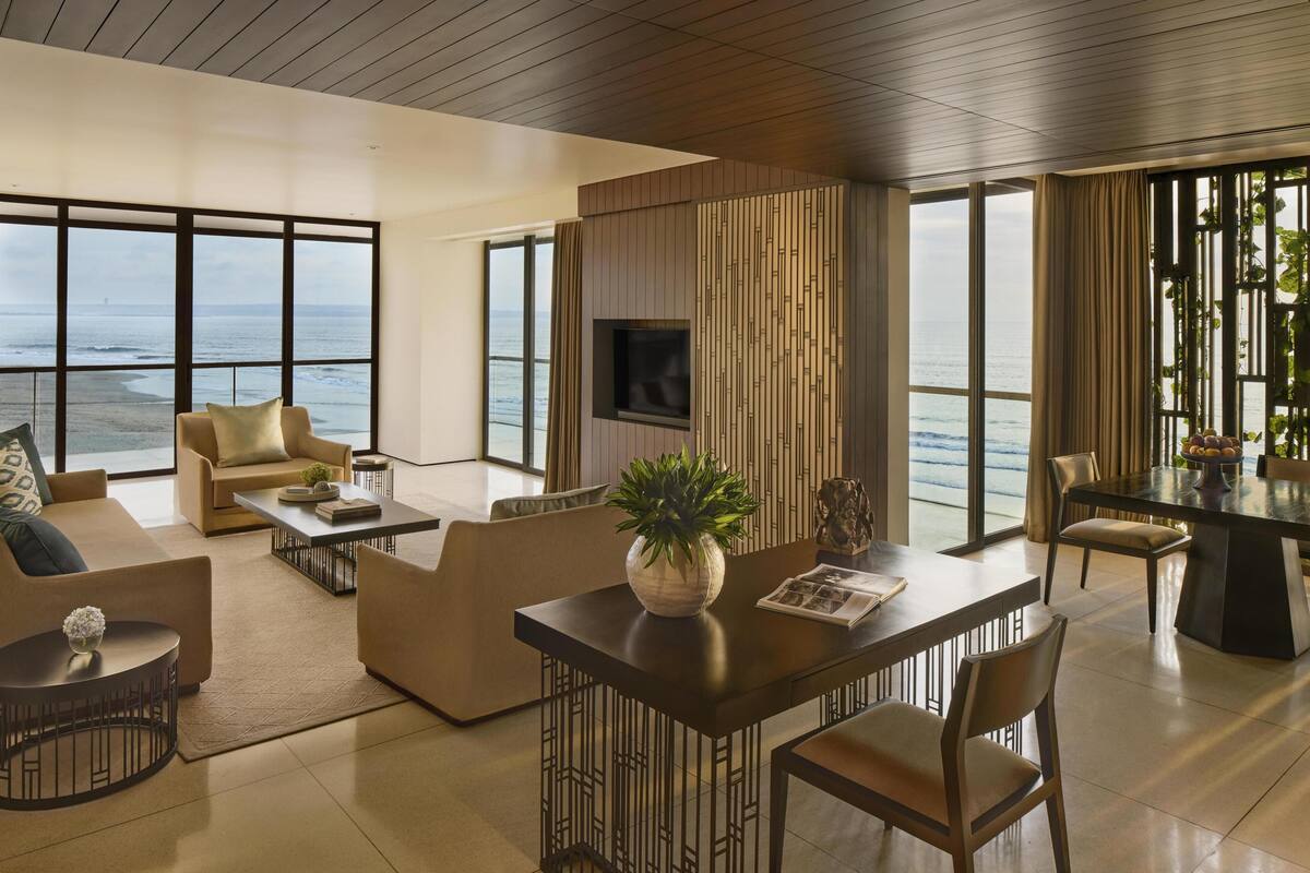 A modern living room with a large window overlooking the ocean, a flat-screen TV mounted on a wooden panel, and a dining area with a table and chairs.