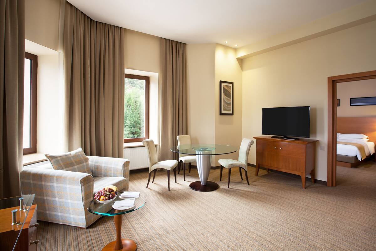 A hotel room with a sofa, a glass-top dining table, a TV, a bed, and a window with curtains.