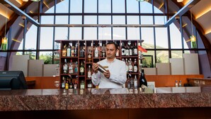 A bar area with a bartender preparing a drink, shelves of liquor behind him, and a large window with a view of the outdoors. - Grand Resort Jermuk (Jermuk)