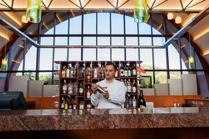 Un espace bar avec un barman préparant un cocktail, des étagères remplies de bouteilles d’alcool derrière lui et une grande fenêtre donnant sur l’extérieur.