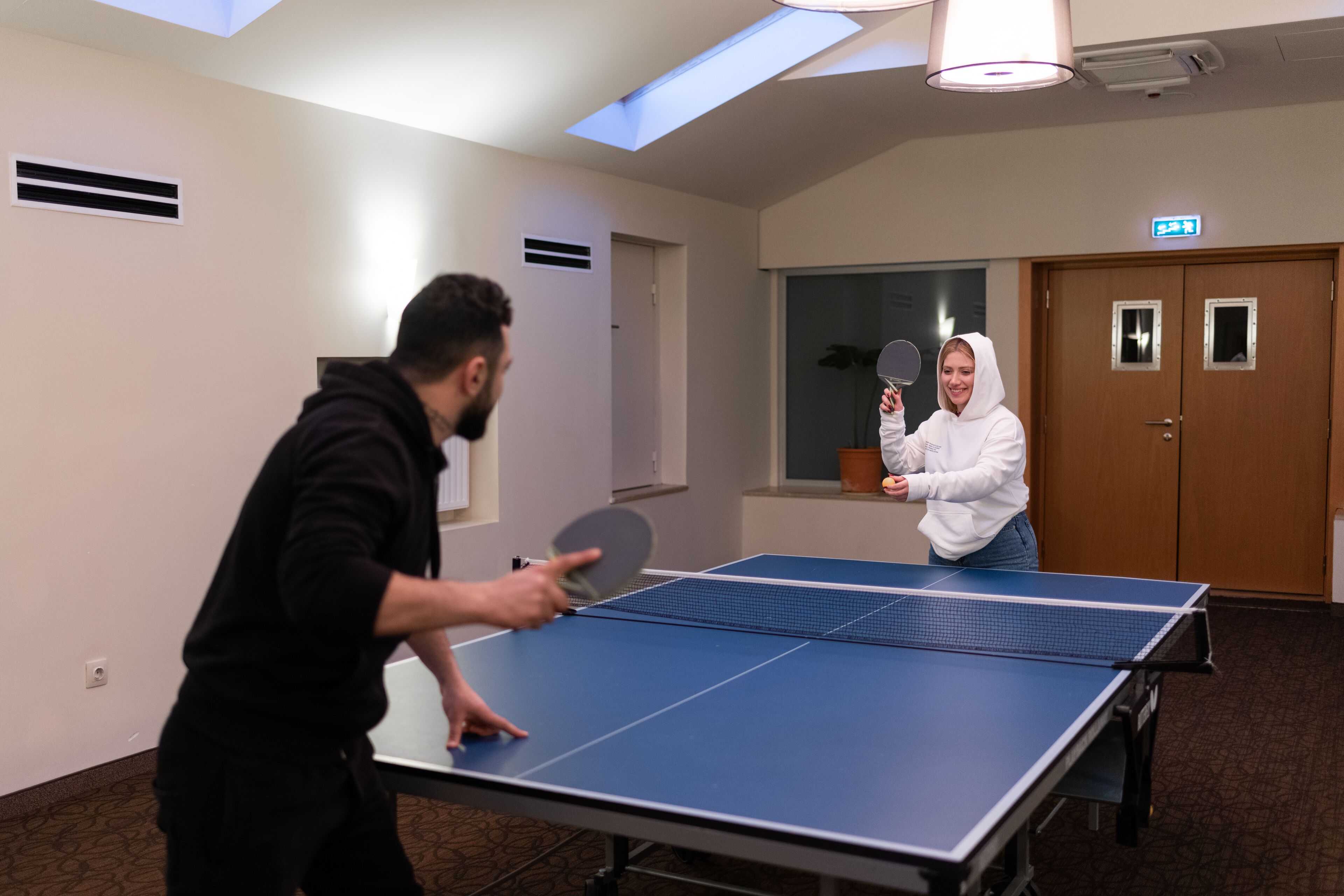 A room with a ping pong table, a door with an exit sign, and a window with a view.