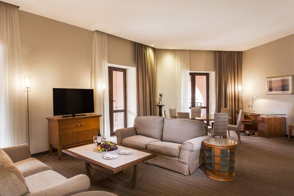 A hotel room with a sofa, a television, a dining table with a fruit bowl, a desk with a chair, and a lamp.