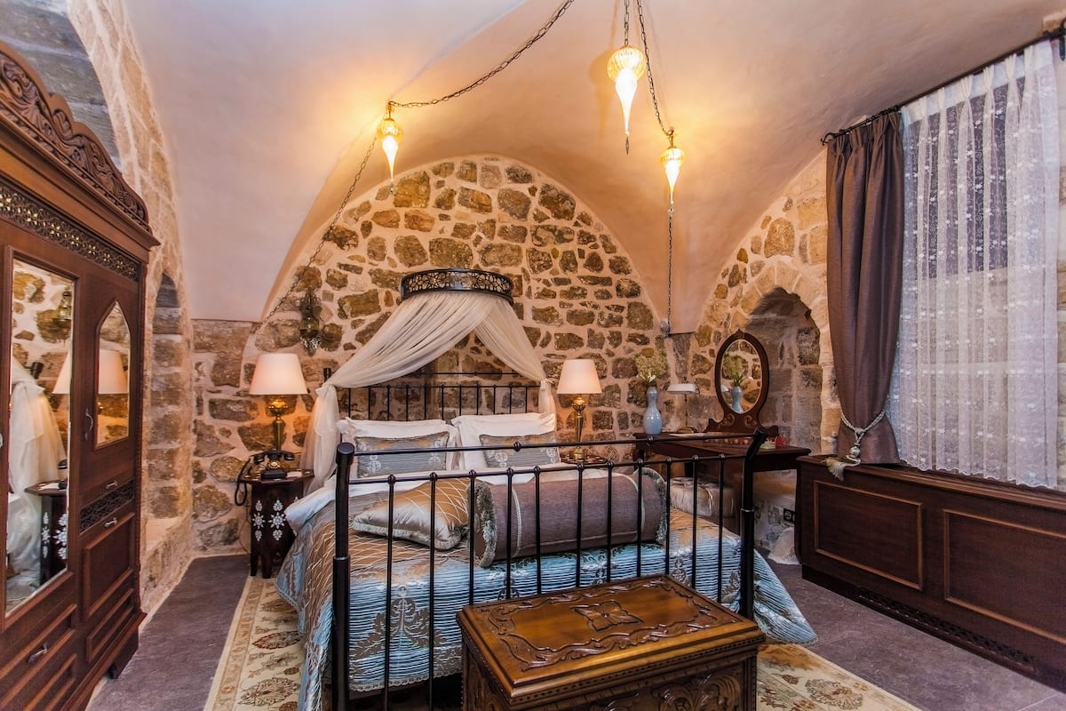 Quarto com parede de pedra, cama de dossel, cômoda de madeira e mesa de cabeceira com abajur.