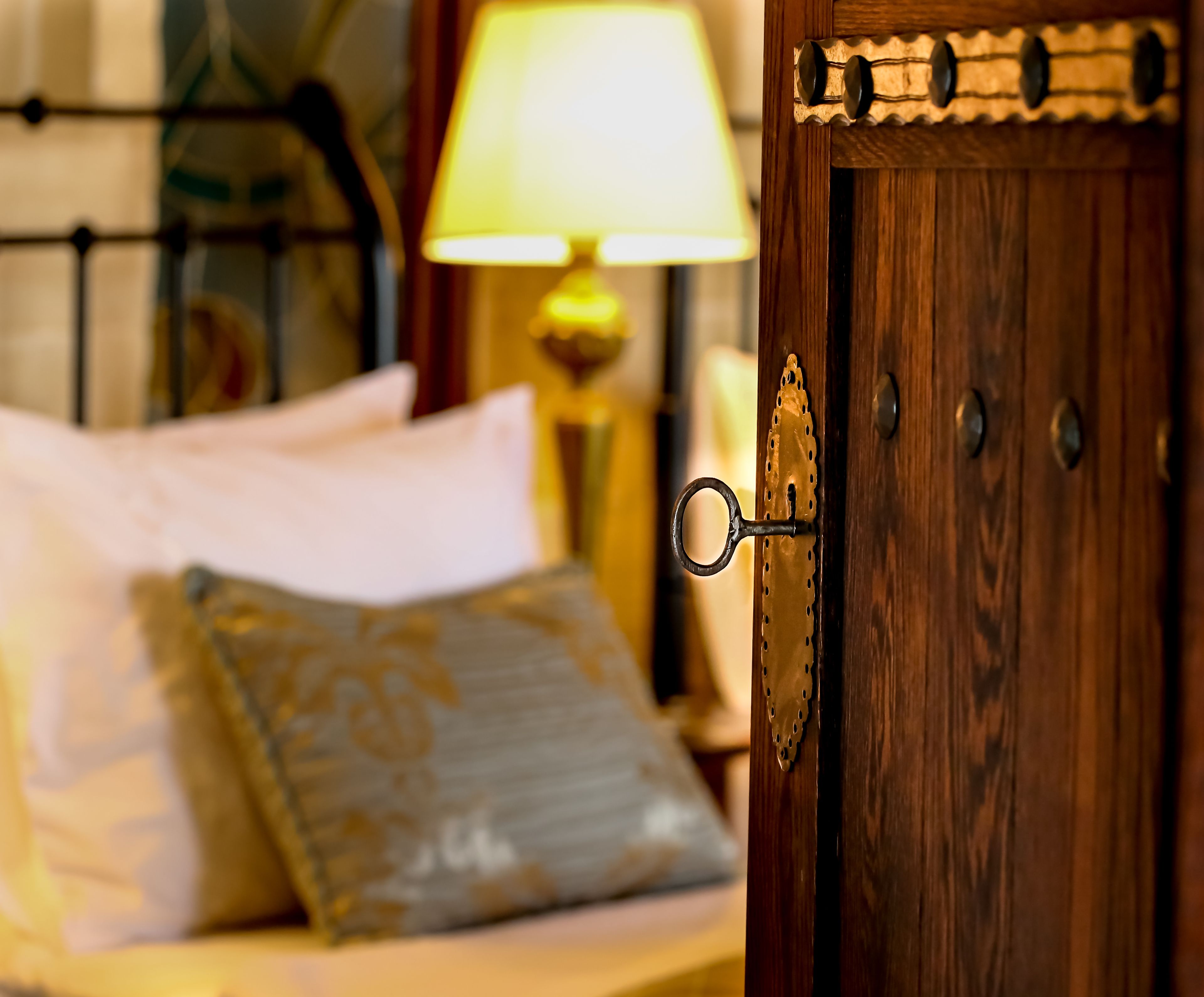 Una puerta de madera con manija de bronce, una cama con almohadas blancas y estampadas, y una lámpara con pantalla amarilla al fondo.