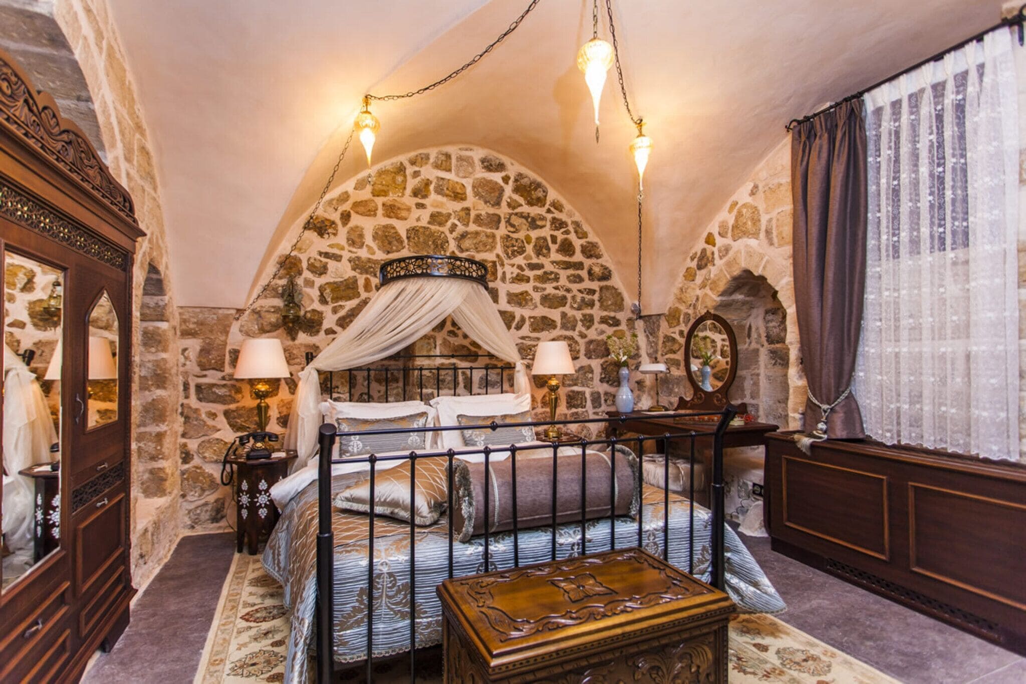 A stone-walled bedroom with a four-poster bed, a nightstand, a dresser, and a mirror.