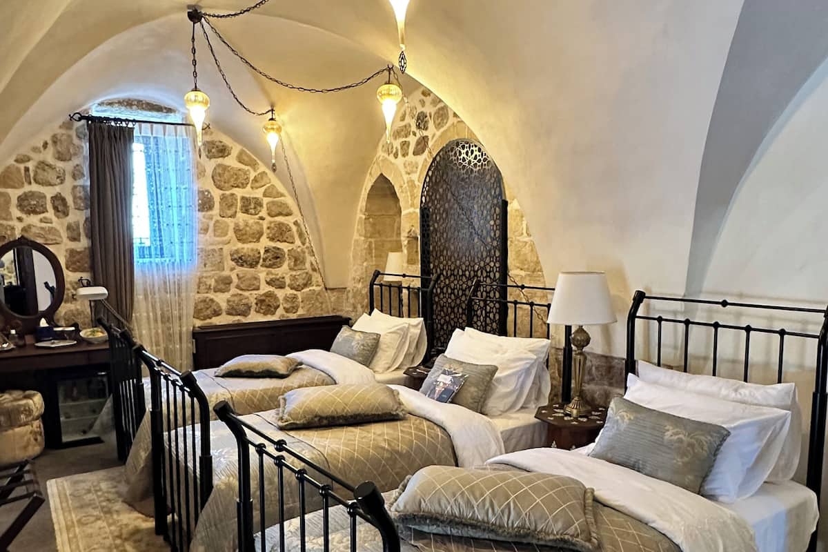 Quarto aconchegante com paredes de pedra, uma cama grande com muitas almofadas, uma escrivaninha com abajur e um espelho decorativo.