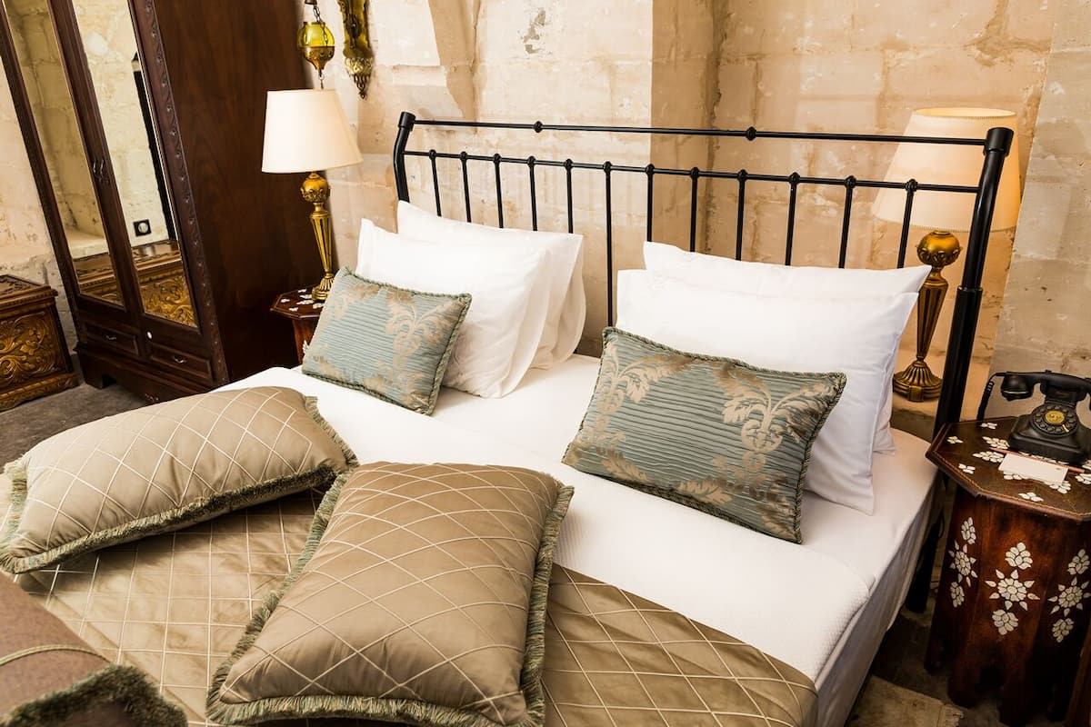 Uma cama bem arrumada com cabeceira de metal, duas almofadas brancas e várias almofadas decorativas. Há duas mesas de cabeceira com luminárias e um telefone antigo.