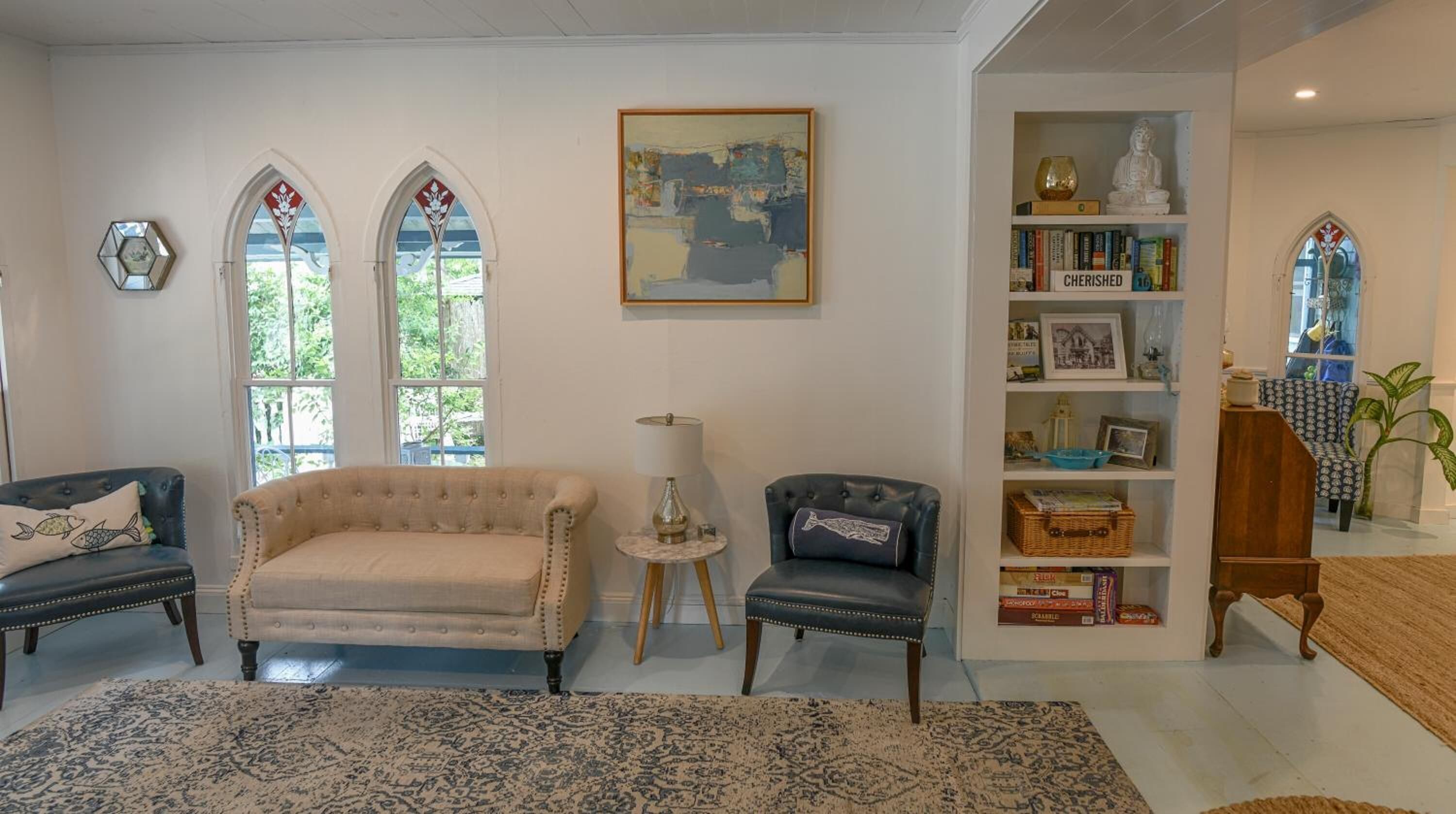 Uma sala de estar com um sofá bege, duas poltronas, uma mesinha com abajur, uma estante de livros e uma estátua.