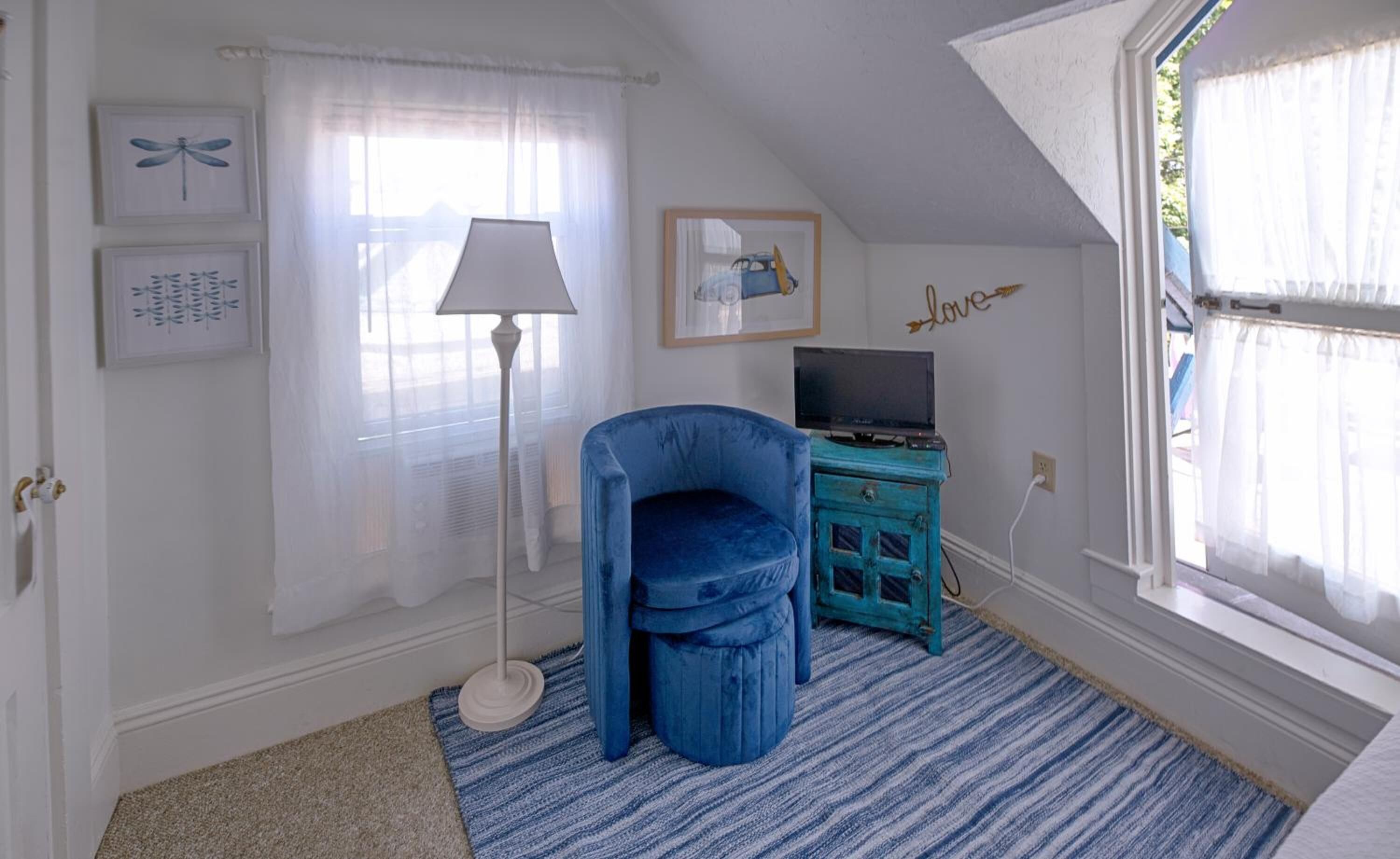 Ein Zimmer mit blauem Sessel, einer Lampe, einem Fernseher auf einem blauen Schrank und einem Fenster mit durchsichtigen Vorhängen.