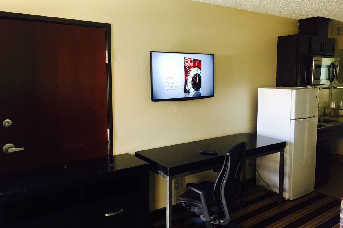 Habitación de hotel pequeña con kitchenette, una mesa de comedor y una silla, y un televisor en la pared que muestra un anuncio deportivo.