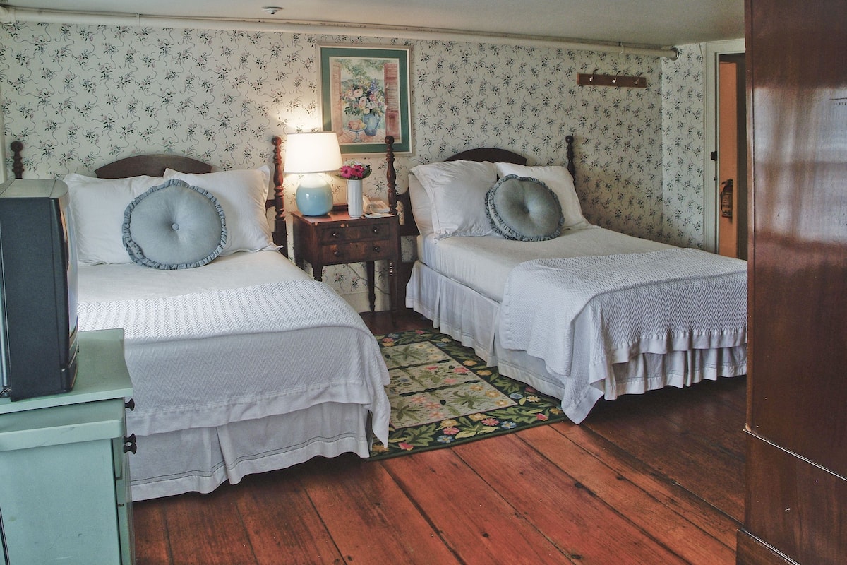 A room with two beds, a nightstand with a lamp, and a floral-patterned wallpaper.