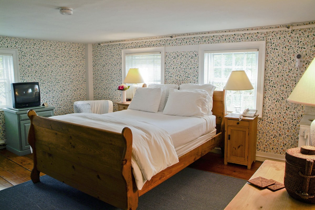 A bedroom with a wooden bed, two bedside tables, a nightstand, a television, and floral wallpaper.