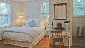 A bedroom with a bed, a desk with a chair, a lamp, a vase with flowers, and two windows with shutters. - Ships Inn (Nantucket)