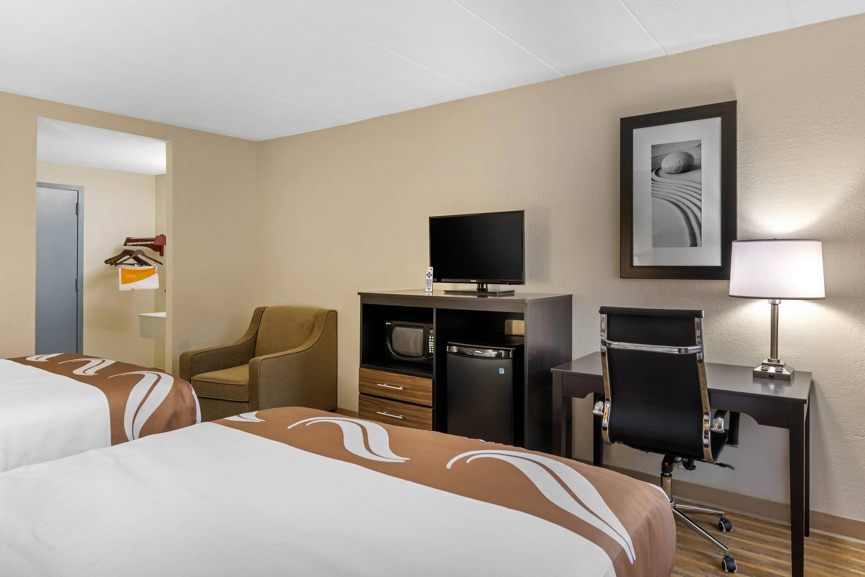 Habitación de hotel con dos camas, un escritorio con ordenador, televisor, una silla y un refrigerador pequeño.