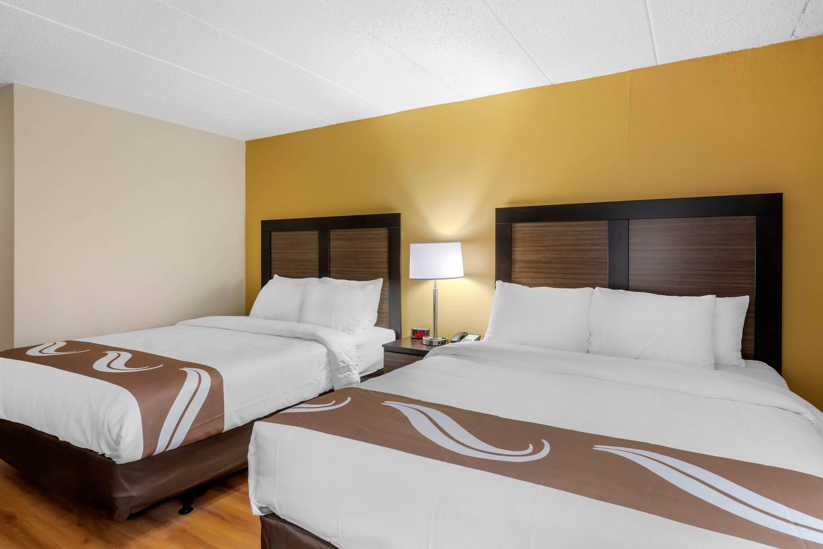 A hotel room with two beds, a nightstand, a lamp, and a clock.