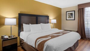 A hotel room with a large bed, two bedside tables, a nightstand, a lamp, a framed picture on the wall, and a window with curtains. - Quality Inn & Suites Ruther Glen (Ruther Glen)