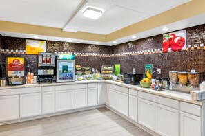 Zona de desayuno autoservicio con máquinas de café, vitrina refrigerada y diversas opciones de alimentos.