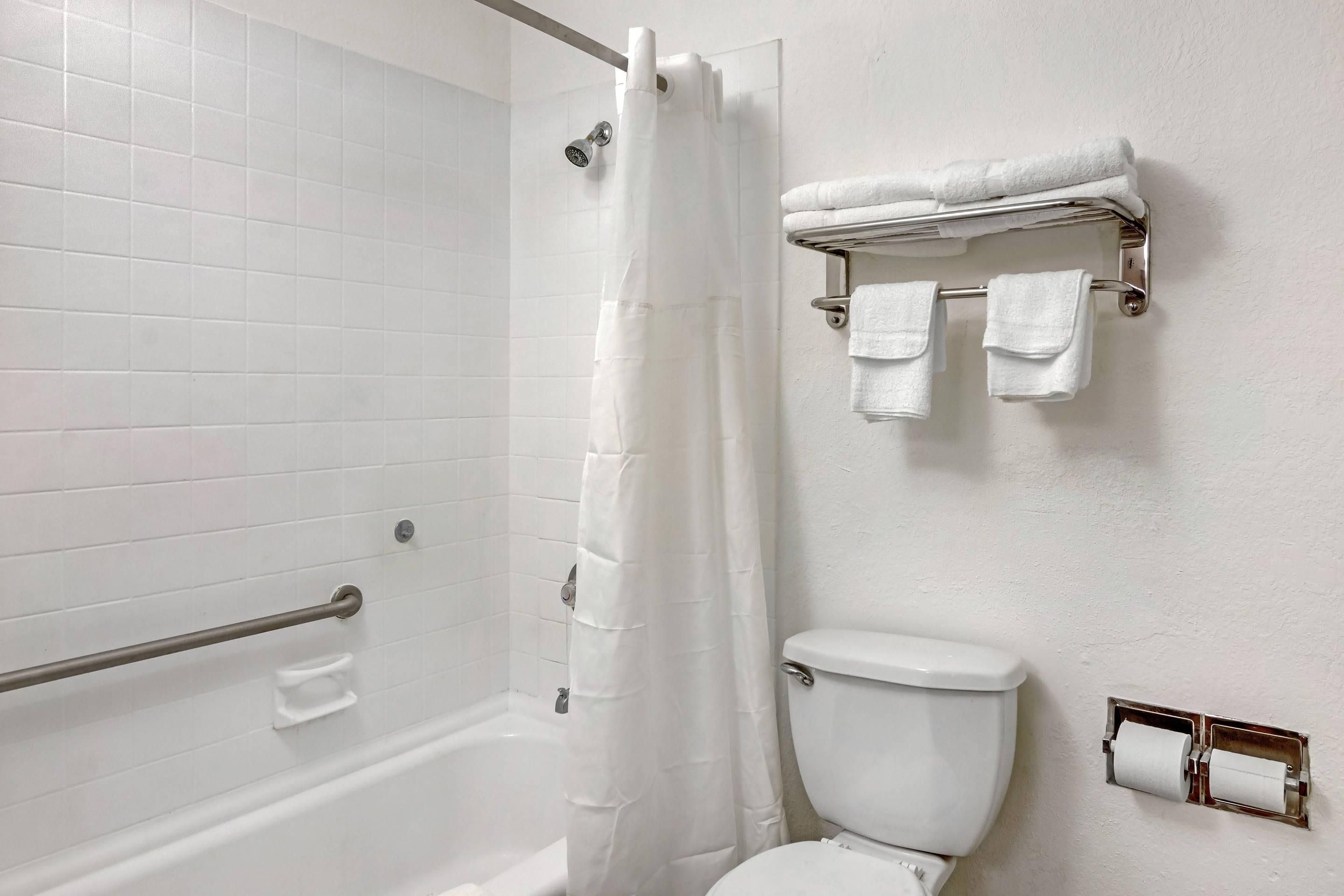 Une salle de bain avec un mur recouvert de carreaux blancs, une douche avec un rideau blanc, une barre d’appui, des toilettes et un porte-serviettes avec deux serviettes.