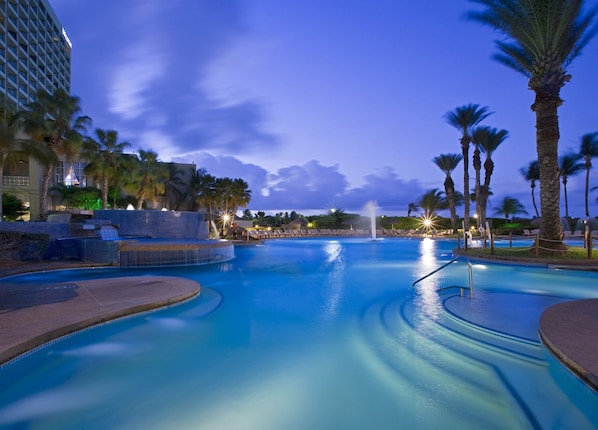 A large swimming pool with a fountain, surrounded by palm trees and lounge chairs, with a hotel building in the background. - Quality Inn & Suites Ruther Glen (Ruther Glen)