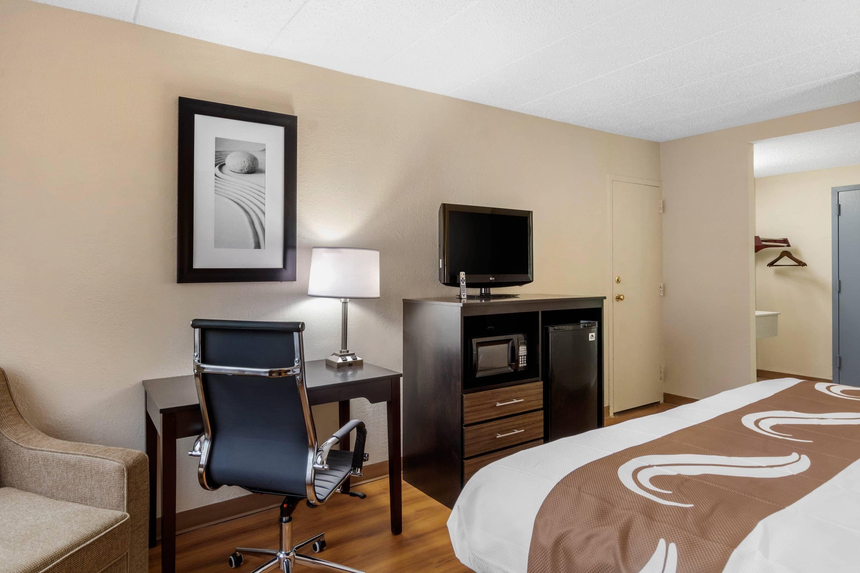 A hotel room with a bed, a desk with a chair, a television, a microwave, and a framed picture on the wall.