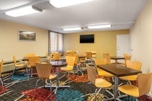 A hotel room with a dining area featuring round tables and chairs, a flat-screen TV mounted on the wall, and a door on the right.