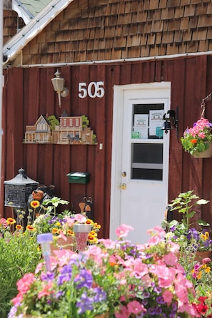 Un bâtiment en bois avec le numéro 505 sur la porte, une petite maquette de maison au-dessus de la porte et un panier à fleurs suspendu.