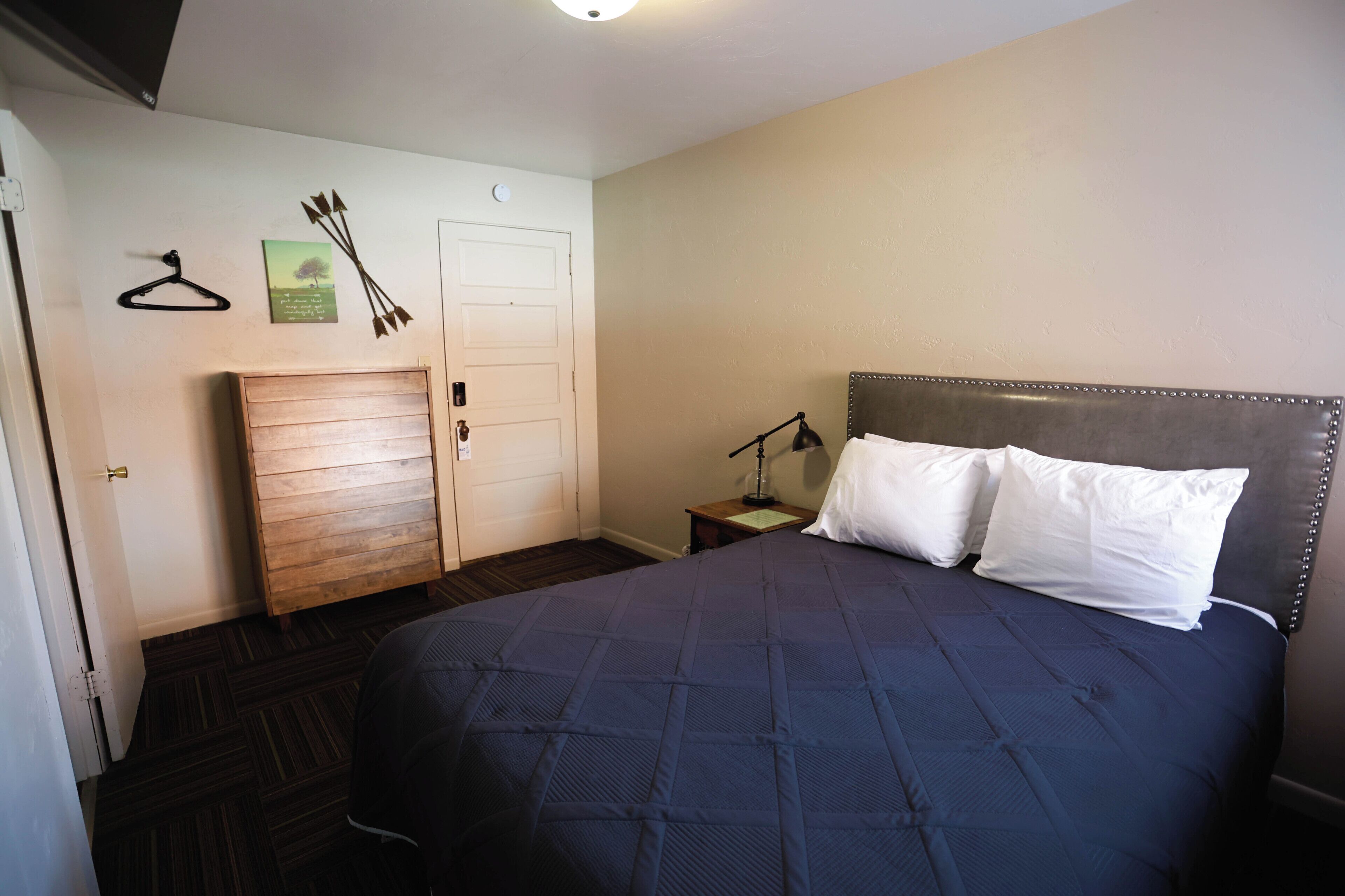 A bedroom with a bed, a nightstand, a hanging light, a door, and a coat rack.