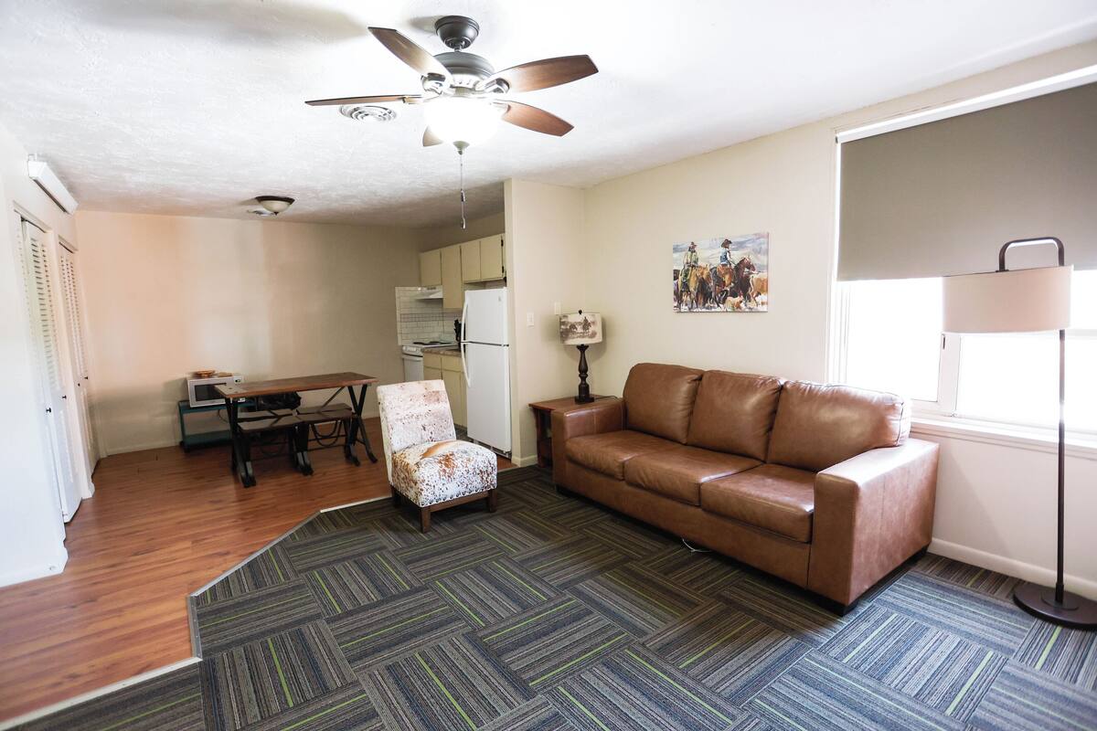 A living room with a brown leather sofa, a patterned carpet, a ceiling fan, and a dining area with a table and chairs.