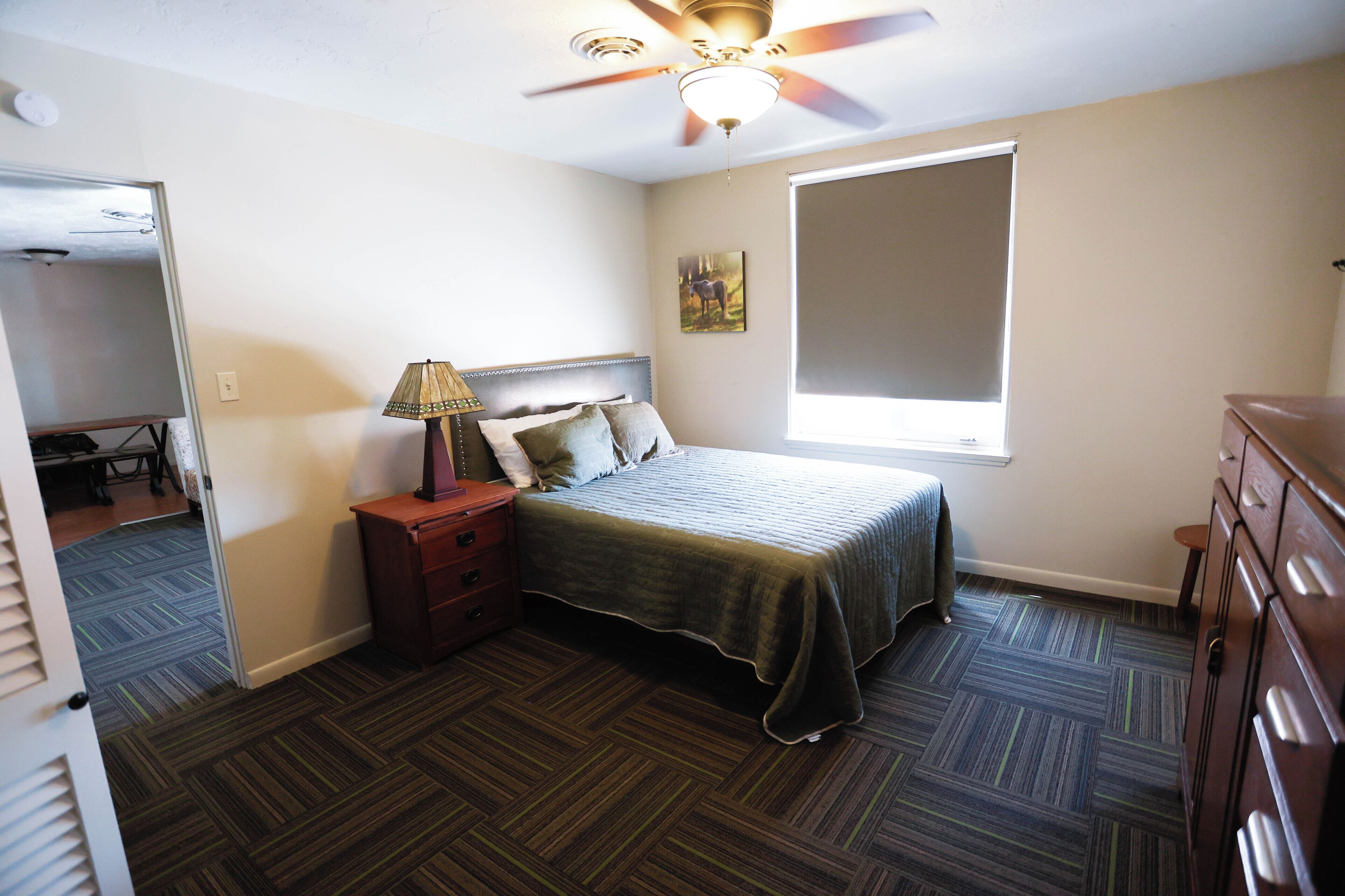 A bedroom with a bed, a nightstand, a lamp, a dresser, a ceiling fan, and a window.
