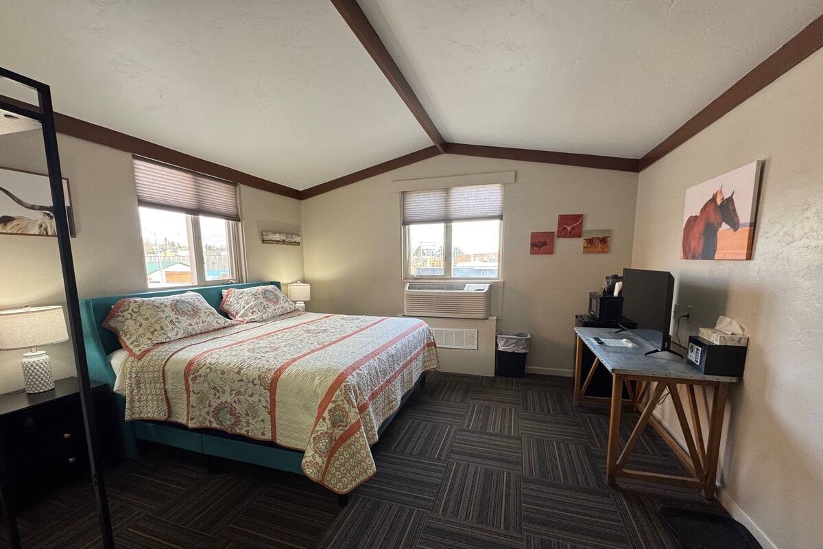 A bedroom with a bed, a desk, a chair, a TV, and a window.