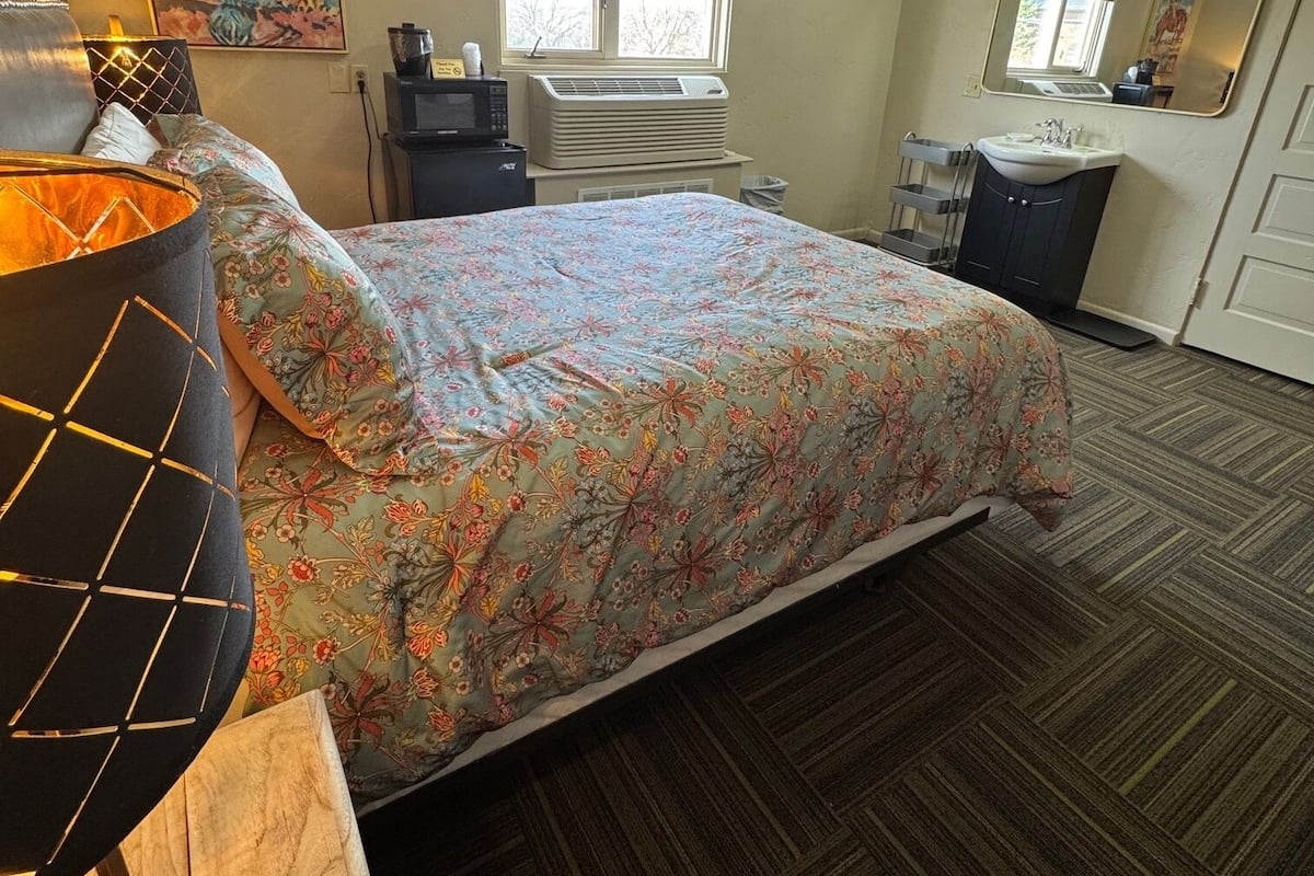A bedroom with a bed, a desk with a lamp, a window with blinds, an air conditioning unit, a sink, and a mirror.