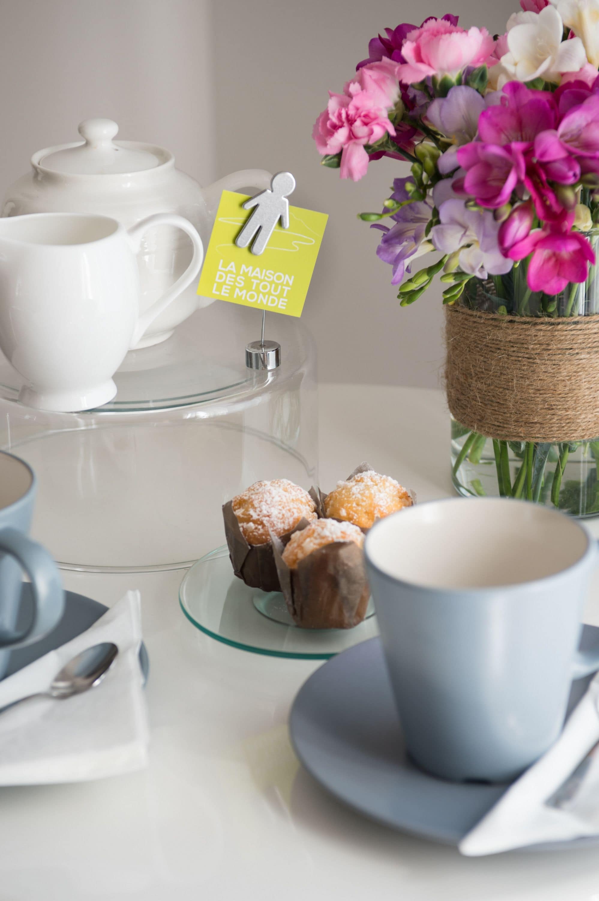 A table with a teapot, cups, a glass cake stand with pastries, and a vase with flowers.