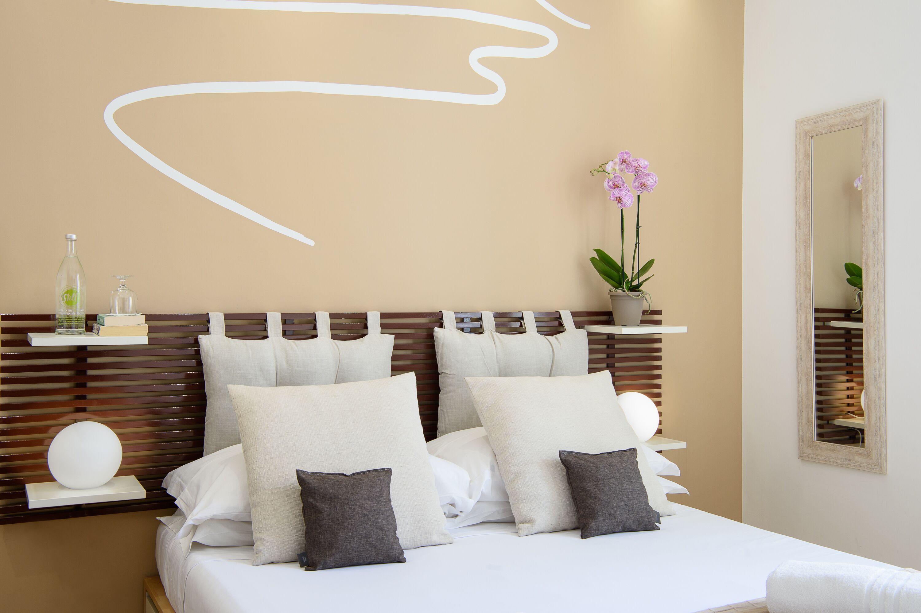 A neatly made bed with white and gray pillows, a bedside table with a bottle and books, and a wall with a mirror and a potted orchid.