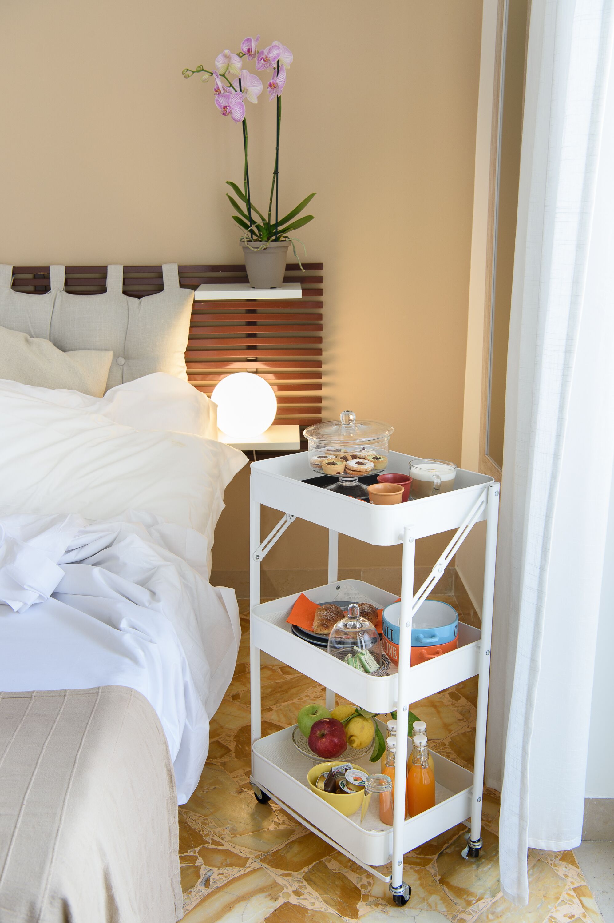 Una cama bien hecha con sábanas blancas, una mesita de noche con una lámpara, un carrito blanco pequeño con productos para el desayuno y una orquídea en maceta sobre la mesita.