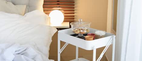 A neatly made bed with white linens, a bedside table with a lamp, a small white cart with breakfast items, and a potted orchid on the nightstand.