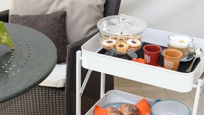 Une chaise en osier avec un coussin, une table avec une assiette verte et un chariot blanc contenant de la nourriture et des boissons.