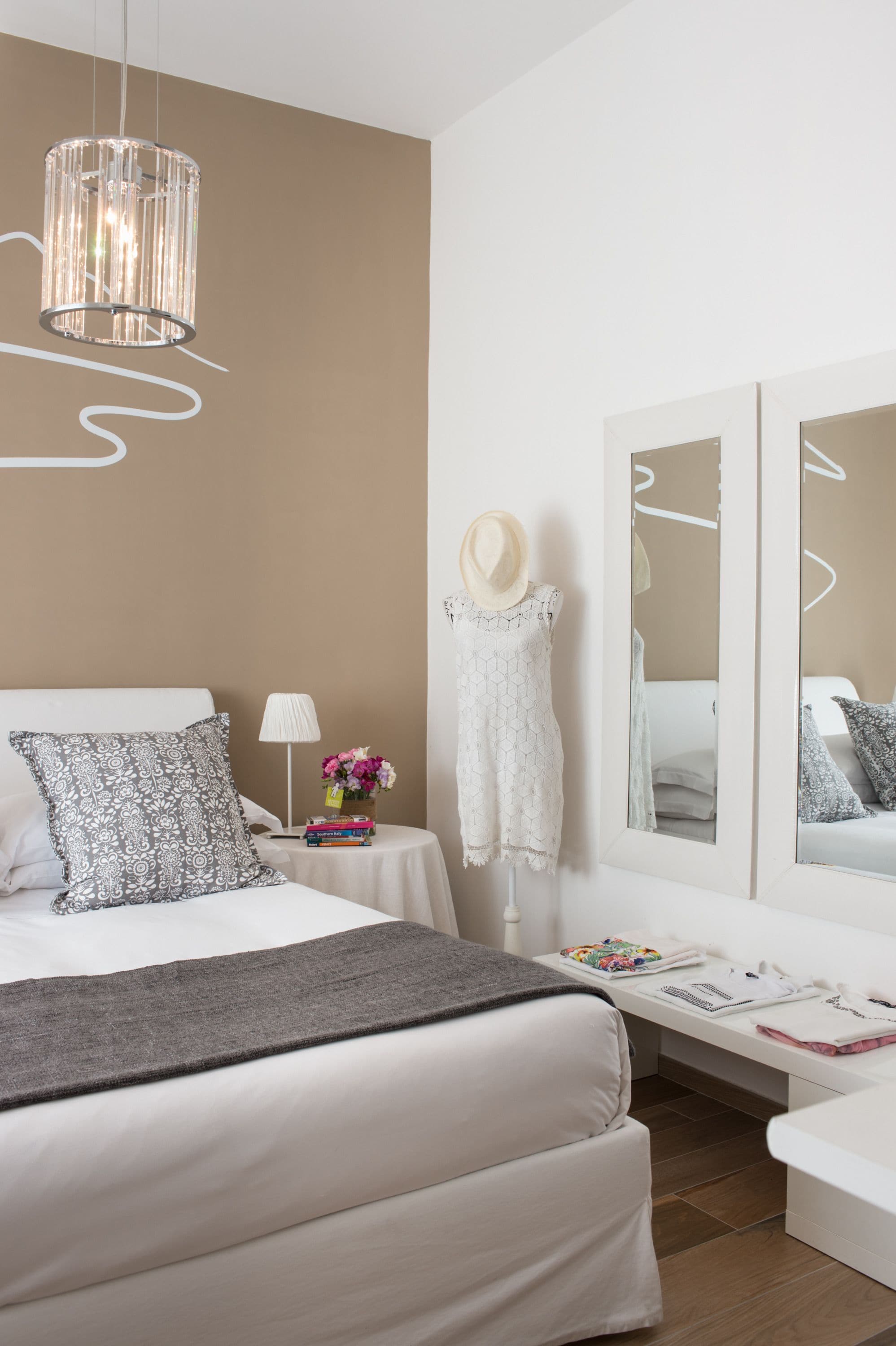 A modern bedroom with a large bed, a bedside table with a lamp and books, a dresser, and a mirror.