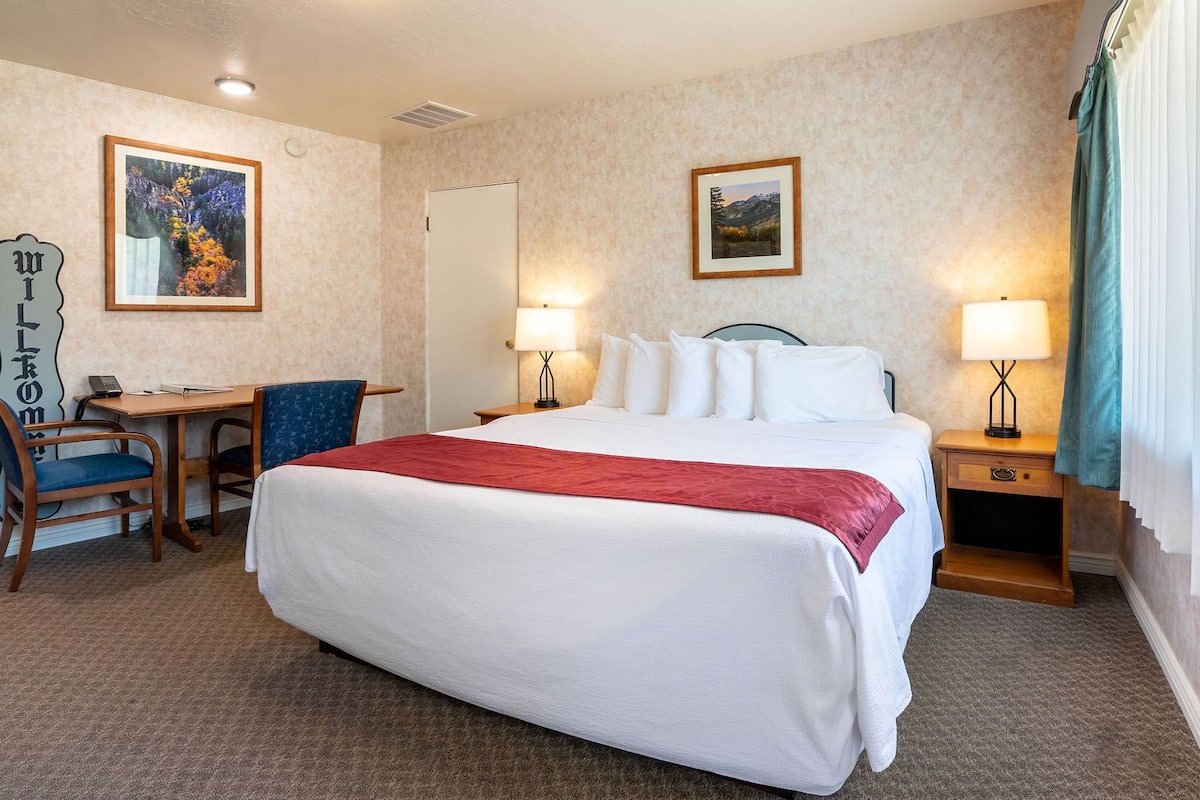 A hotel room with a large bed, two bedside tables with lamps, a desk with a chair, and two framed pictures on the walls.