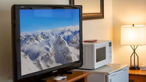 En tv-apparat står på ett träställ och visar ett snöigt berglandskap. Bredvid står en mikrovågsugn och på ett nattduksbord finns en lampa.