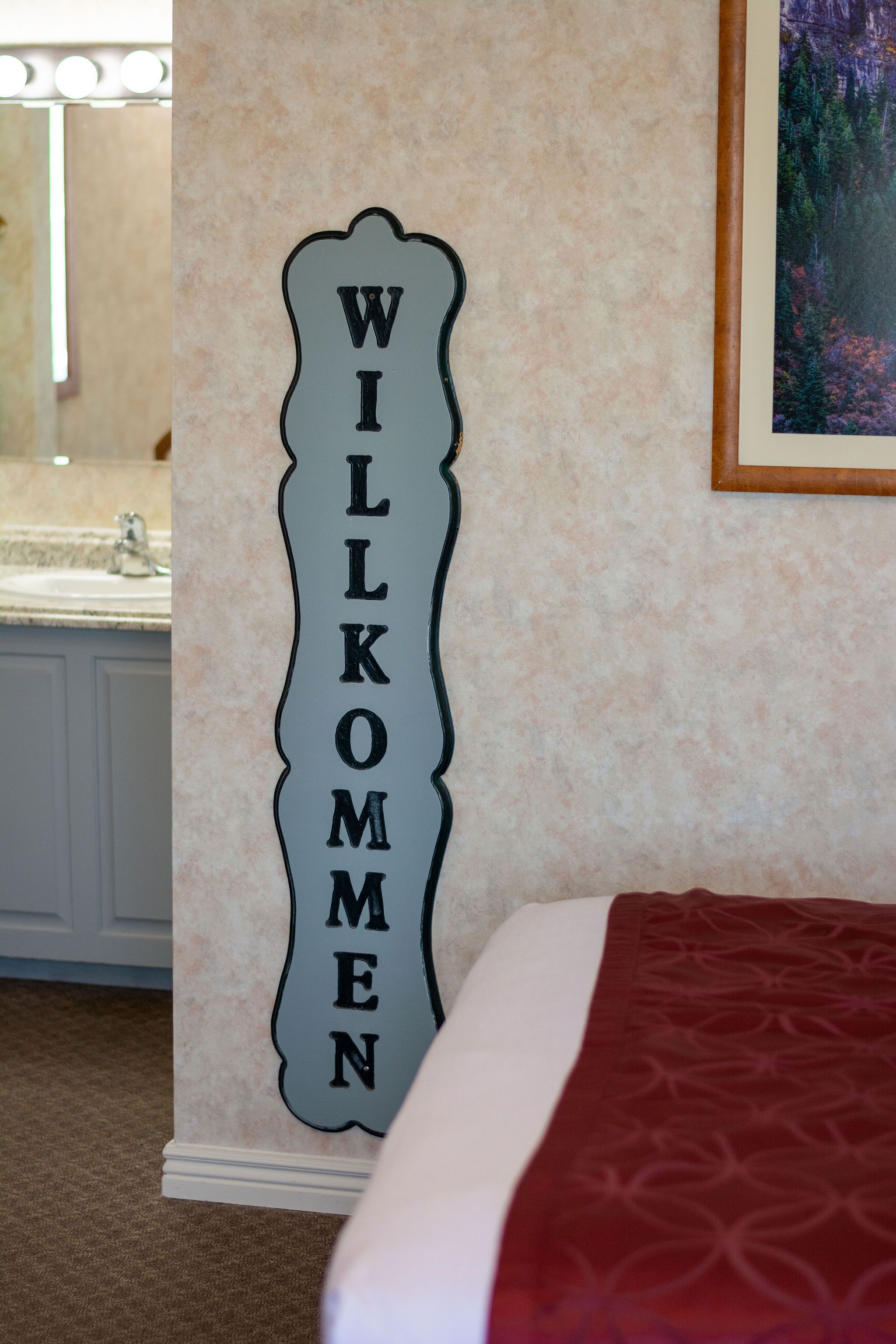 A wall-mounted sign with the text 'WILKOMEN' in a decorative font, a bathroom with a sink and mirror, and a bed with a patterned bedspread.