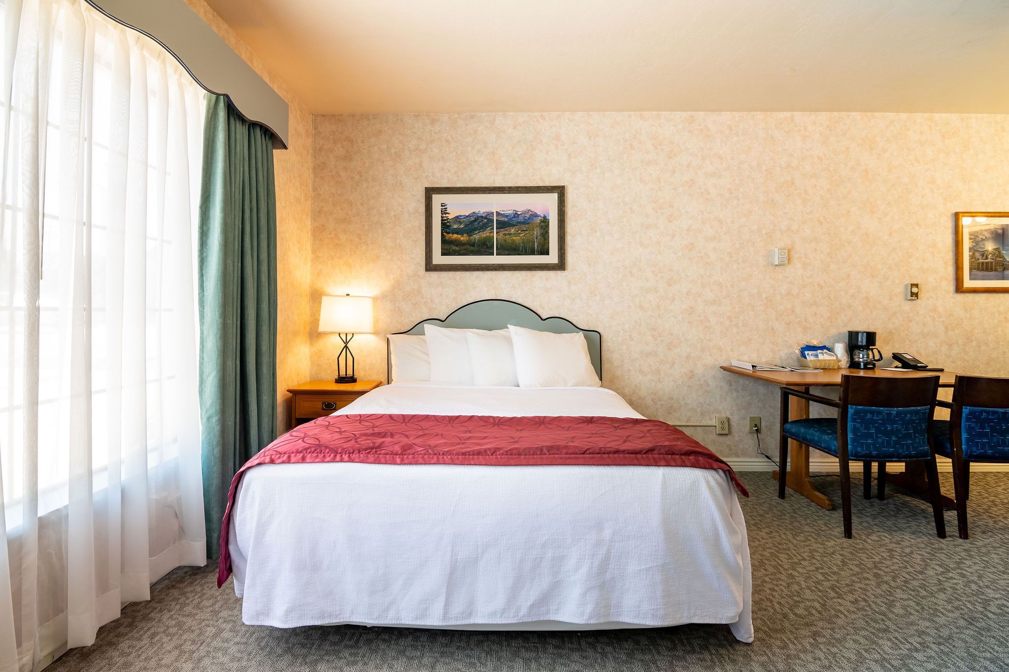 A hotel room with a bed, a desk, chairs, a lamp, and a framed picture on the wall.