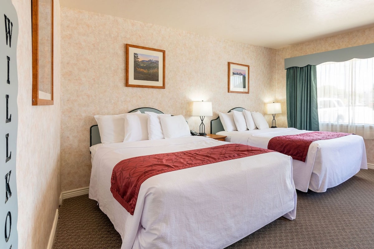A hotel room with two beds, a large window with curtains, and two framed pictures on the wall.
