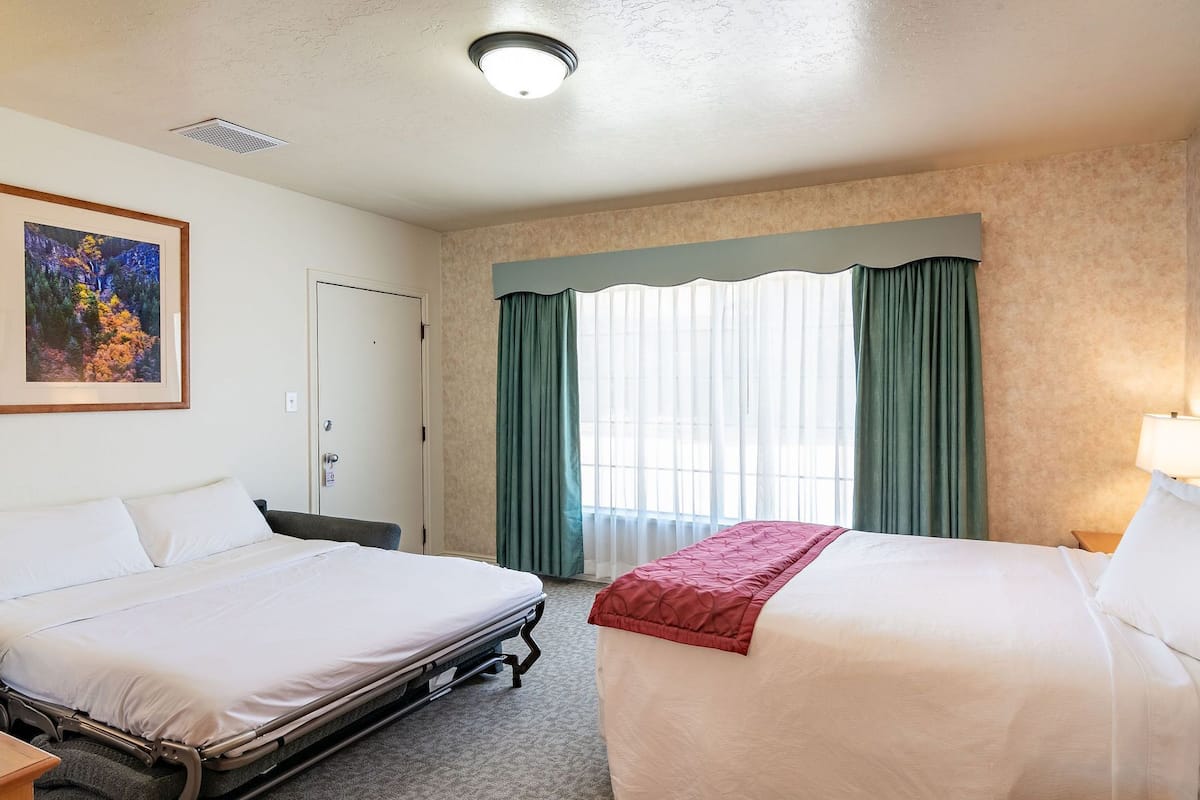 A hotel room with two beds, a desk, a chair, a window with curtains, and a framed picture on the wall.